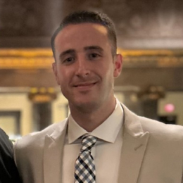 A man in a light-colored suit and checkered tie stands indoors, smiling slightly, with a blurred ornate background behind him at FutureCon OC 2024.
